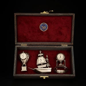 May include: A dark wooden display box with a deep red velvet interior containing three silver-toned metal desk clocks. The clocks are shaped like a hot air balloon, a sailing ship, and a bell. A blue circular label reads Nazfan Antique Gallery.