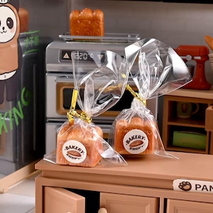May include: Two miniature loaves of bread packaged in clear plastic bags with gold twist ties. Each bag features a round white sticker with the text BAKERY SHOP and an illustration of a loaf of bread. The items are placed on a miniature kitchen counter.