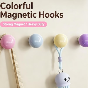 May include: Four spherical magnetic hooks in pink, light blue, yellow, and purple. The text "Colorful Magnetic Hooks" and "Strong Magnet / Heavy Duty" appears above the hooks. A beige strap hangs from the pink hook, and a light blue jellyfish charm hangs from the yellow hook.
