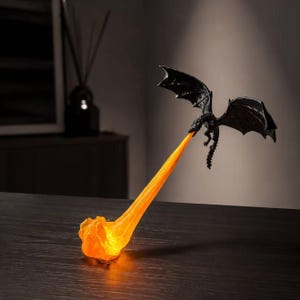 May include: A black dragon figurine in flight, suspended in the air, emitting a long, translucent orange flame that extends downward to touch the surface. The dragon features detailed wings and a textured body, with the light glowing from the flame.