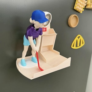 Puede incluir: Un organizador de pared magnético impreso en 3D con una figura que lleva una gorra azul, una camiseta morada y pantalones cortos azul claro. La figura sostiene un palo de golf rojo sobre una plataforma beige con un pequeño compartimento marcado con el número 19.