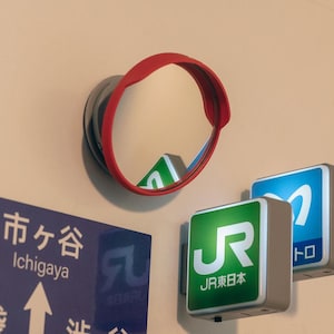 May include: A circular convex mirror with a red frame mounted on a wall. Below it are two illuminated square signs, one green with the white JR East logo and one blue with a white logo, alongside a blue sign with the text Ichigaya and an arrow.