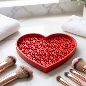 Op de afbeelding: Een hartvormige rode siliconen make-up kwastenreinigingsmat met een getextureerd oppervlak van vele kleine verhoogde hartpatronen, omringd door diverse make-up kwasten met roségouden metalen hulzen en zachte haren.