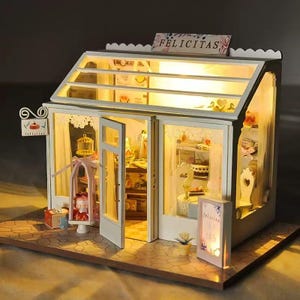 May include: A miniature white bakery model with a sloped glass roof, illuminated interior, and an open door. The sign on the roof reads FELICITAS. The shop features a display stand with a cake, a small sign, and detailed interior furnishings.