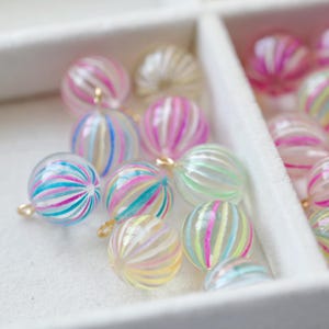 Könnte beinhalten: Eine Sammlung kugelförmiger, klarer Glasperlen mit innenliegenden vertikalen Streifen in verschiedenen Farben wie Pink, Blau, Gelb und Grün. Jede Perle hat eine kleine goldfarbene Metallöse an der Oberseite zum Aufhängen oder Basteln.