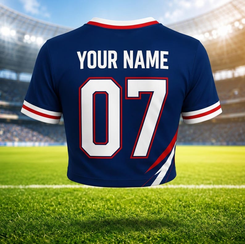 May include: A navy blue sports jersey with the text "YOUR NAME" above the number "07" in white. The jersey has red and white accents on the sleeves and collar. The background shows a green field and a stadium.
