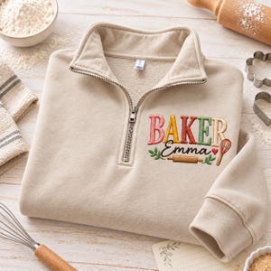 May include: A beige quarter-zip sweatshirt featuring embroidered text that reads BAKER Emma in multi-colored letters. The design includes a small red heart, a brown rolling pin, green leaves, and a red whisk icon below the name.
