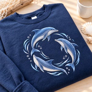 Peut inclure: Un sweat-shirt bleu marine à col rond orné d'une broderie représentant trois dauphins nageant en cercle. Les dauphins sont déclinés dans des tons de bleu, gris et blanc, entourés de vagues et de bulles d'eau stylisées brodées sur le tissu.