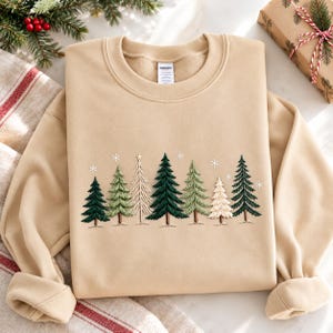 May include: A beige crewneck sweatshirt featuring an embroidered design of various pine trees in shades of dark green, light green, and cream, accented with small white embroidered snowflakes scattered above the tree line.