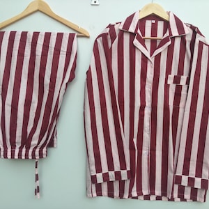 May include: A pajama set consisting of a long-sleeved button-down shirt with a collar and a chest pocket, and matching pants with an elastic waistband and drawstring. Both pieces feature a vertical striped pattern in dark red and light pink.