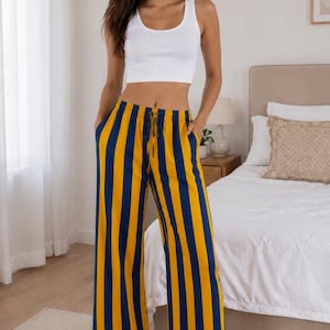 Op de afbeelding: Een wit mouwloos crop topje gecombineerd met een wijde loungebroek met verticale strepen in mosterdgeel en marineblauw. De broek heeft een elastische tailleband met een trekkoord en twee zijzakken, en valt tot op de grond.