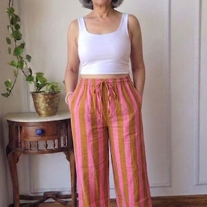 May include: A white sleeveless vest top paired with wide-leg trousers featuring vertical stripes in alternating shades of brown and pink. The trousers have an elasticated waistband with a drawstring tie and two side pockets, ending at the ankles.