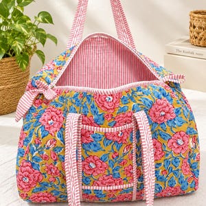 Könnte beinhalten: Eine gesteppte Reisetasche mit einem lebendigen Blumenmuster aus rosa Blüten und blauen Blättern auf senfgelbem Grund. Die Tasche verfügt über pink-weiß gestreifte Griffe, Kanten und ein Innenfutter im passenden Streifendesign.
