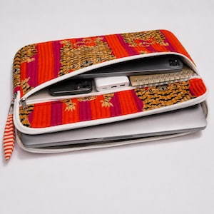 May include: A quilted laptop sleeve featuring a vibrant orange and magenta striped pattern with tiger motifs. The sleeve has two zippered compartments containing a silver laptop, a black smartphone, a white power bank, and a spiral notebook.
