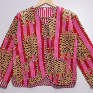 Puede incluir: Una chaqueta acolchada con un patrón vibrante de tigres sobre un fondo de rayas verticales rosas y rojas. La chaqueta tiene un cuello en V, mangas largas con puños doblados y dos bolsillos delanteros. El forro interior es de rayas.