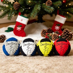 May include: Four colourful Dirtbag balls are displayed in front of a Christmas tree with stockings and pine cones. The balls are blue, black, yellow, and red, each with a black top and the Dirtbag logo. The balls are approximately 10 cm in diameter.