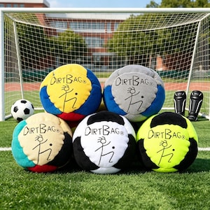 Puede incluir: Una colección de balones de fútbol coloridos con el texto "DIRTBAG" y un logotipo de figura de palo. Los balones son de varios colores, incluyendo amarillo, azul, gris, negro y verde. Una portería de fútbol, un balón de fútbol y espinilleras están en el fondo.