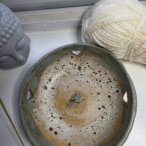 May include: A round, hand-formed ceramic bowl with a speckled beige and brown interior featuring a small heart-shaped cutout at the centre and three additional heart-shaped cutouts along the rim, finished with a textured, muted blue-grey exterior.