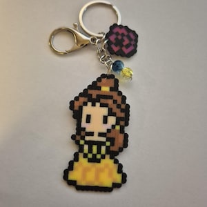May include: A pixel art keychain featuring a yellow dress and brown hair. The keychain includes a silver clasp, a rose charm, and two colorful beads. The design is reminiscent of a princess.