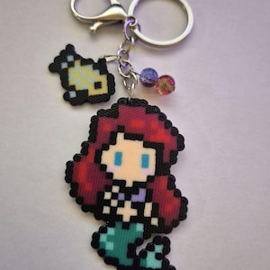 May include: A pixelated keychain featuring a mermaid with red hair, a teal tail, and a small fish. The keychain has a silver clasp and a key ring, with two decorative beads. The mermaid is in a pixel art style.