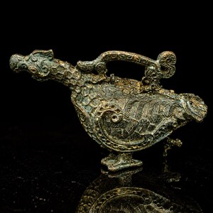 Könnte beinhalten: Ein bronzefarbenes Metallgefäß in Vogelform mit langem Hals und einem geschwungenen Griff auf dem Rücken. Die Oberfläche weist komplexe, erhabene Reliefmuster auf, die an Federn und Schuppen erinnern, mit einem verwitterten, dunklen Patina-Finish.