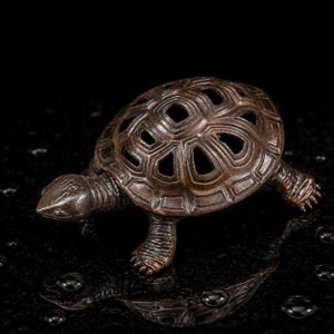 May include: A dark brown, metallic turtle figurine featuring a hollow, open-work shell design with geometric cutouts. The turtle is depicted in a walking stance with textured skin on its head and legs, showcasing a weathered, bronze-like finish.