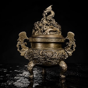 May include: A metallic bronze-toned incense burner featuring intricate dragon relief carvings on the body and lid. The vessel stands on three legs, includes two ornate side handles, and displays a detailed dragon sculpture atop the removable lid.