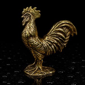 Puede incluir: Una pequeña figura de gallo de color oro metálico en perfil. La escultura presenta plumas texturizadas detalladas en el cuello, las alas y la cola, con una cresta prominente en la cabeza y un pico afilado, todo en un acabado de latón pulido.