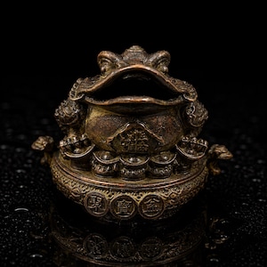 May include: A bronze-toned metal sculpture of a three-legged toad with a wide open mouth. The toad sits atop a circular base decorated with embossed circular coin motifs featuring Chinese characters. The surface has a dark, weathered patina.