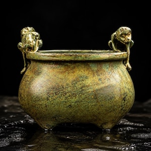 May include: A round, bronze-toned metal vessel with a weathered, mottled green and gold patina. The bowl features two sculpted monkey figures perched on opposite sides of the rim, with small feet extending down the sides of the rounded body.