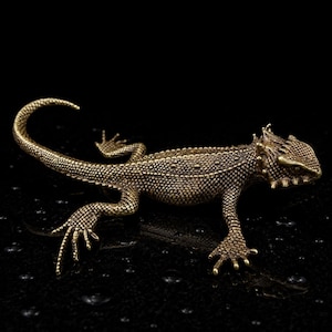 May include: A metallic bronze-toned lizard figurine featuring a highly detailed, textured, scaly surface. The lizard is depicted in a crawling position with its long, curved tail extended and sharp, pointed spikes along its head and back.