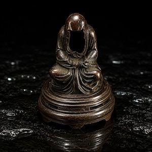 May include: A dark brown, metallic sculpture of a hooded, seated figure with draped robes, positioned on a circular, tiered pedestal base. The figure has a smooth, rounded head covering and folded arms, resting on a matching, ornate, circular stand.