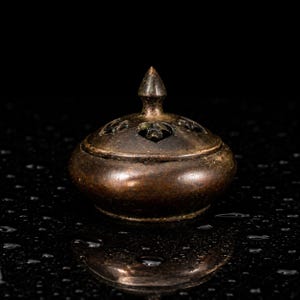 May include: A dark bronze-toned metal incense burner with a rounded base and a removable lid. The lid features a pointed finial at the center and multiple heart-shaped cutouts arranged in a circular pattern to allow for smoke ventilation.