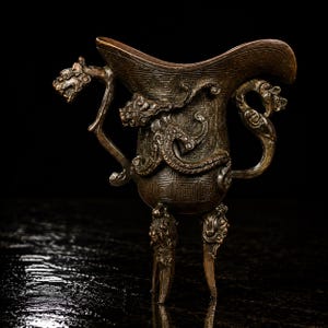 May include: A dark bronze-toned metal vessel featuring a flared rim, three tapered legs, and a handle shaped like a dragon head. The body is decorated with an intricate relief pattern of a coiled dragon and geometric square motifs in a dark finish.