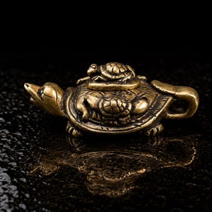 Pode incluir: Uma estatueta de metal cor de latão em forma de uma tartaruga grande com uma tartaruga menor sobre o casco. A superfície tem um brilho metálico com detalhes escuros e reentrantes que destacam os padrões texturizados dos cascos e da cabeça.