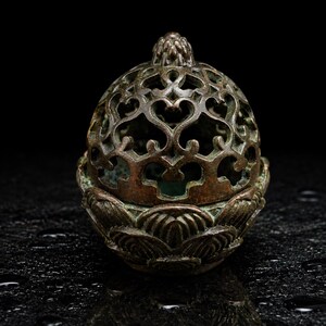 May include: A bronze-toned metal incense burner featuring a dome-shaped lid with intricate openwork heart-shaped cutouts and a finial at the top. The base is designed with layered, sculpted petal motifs resembling a blooming lotus flower.