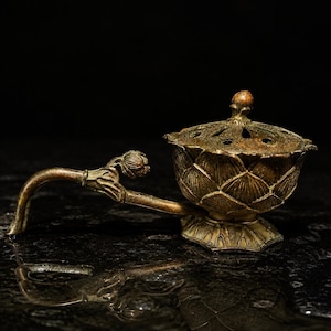 May include: A bronze-toned metal incense burner shaped like a lotus flower with a matching lid featuring cut-out details. The vessel has a long, curved handle that terminates in a small, closed lotus bud, all resting on a wide, petal-shaped base.