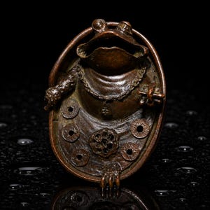 May include: A dark bronze-toned sculpture of a frog with a wide mouth, wearing a chain necklace with a small pendant. The frog is positioned within an oval frame, surrounded by several small circular coin-like discs and a central floral motif.
