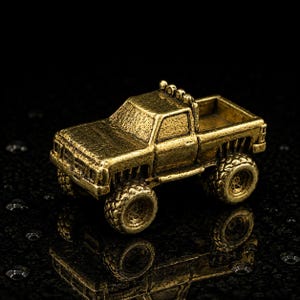 May include: A small, metallic gold-toned pickup truck figurine with a textured finish. The miniature vehicle features a cab, an open truck bed, and four large, rugged wheels. The design includes a roll bar with four circular lights on top.