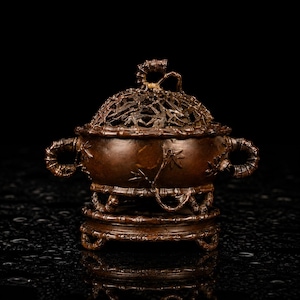 May include: A dark brown bronze incense burner featuring a bamboo-inspired design. The vessel has two side handles, a removable lid with an intricate openwork pattern, and a matching circular base, all decorated with embossed bamboo stalks and leaves.
