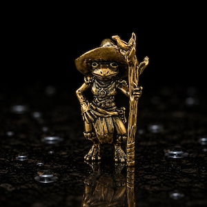 Può includere: Una piccola statuetta in metallo color oro raffigurante una rana in posizione eretta. La rana indossa un cappello a tesa larga e abiti drappeggiati, e tiene un alto bastone nodoso nella mano sinistra. La superficie ha una finitura lucida.