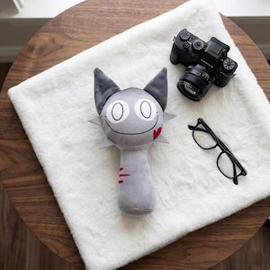 Può includere: Un giocattolo a forma di gatto in peluche grigio con grandi occhi, una bocca sorridente e un dettaglio a forma di cuore rosso. Il giocattolo è su una superficie bianca e soffice, accanto a una fotocamera nera e occhiali.