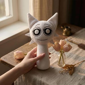 Könnte beinhalten: Ein weißes Plüschtier in Form einer Katze mit großen schwarzen Augen und einem lächelnden Gesicht. Die Katze hat spitze Ohren und sternförmige Details am Kopf. Das Spielzeug wird in einer Hand in der Nähe einer kleinen Vase mit rosa Tulpen gehalten.