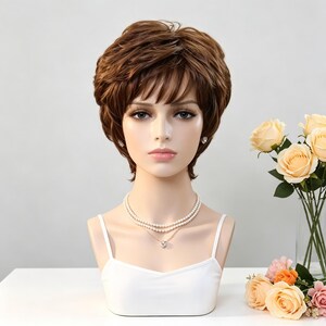 May include: A short, layered brown wig with soft bangs styled on a mannequin head. The mannequin wears a double-strand pearl necklace with a small pendant and a white camisole top. The hair features natural highlights and a textured finish.