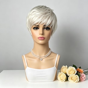 May include: A mannequin head wearing a short, layered, platinum blonde wig with side-swept bangs. The mannequin features a smooth, light-toned complexion, defined eyebrows, and pink lips. It is adorned with a double-strand pearl necklace and a white top.