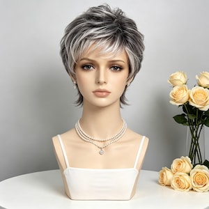 Peut inclure: Une perruque courte et dégradée composée d'un mélange de mèches gris foncé et blanc éclatant. La perruque est présentée sur une tête de mannequin portant un collier de perles à plusieurs rangs avec un petit pendentif et un caraco blanc.