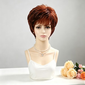 May include: A short, layered auburn wig with side-swept bangs styled on a mannequin head. The mannequin wears a white camisole and a double-strand pearl necklace. The hair features deep reddish-brown tones with subtle highlights and volume.