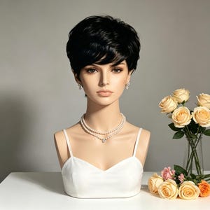 May include: A mannequin head wearing a short, layered black wig with side-swept bangs. The mannequin features a white camisole, a double-strand pearl necklace, and small pearl stud earrings. The facial features are neutral with light skin tone.