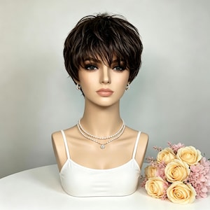 May include: A mannequin head wearing a short, layered dark brown wig with textured bangs. The mannequin features a white camisole, a double-strand pearl necklace with a central pendant, and small pearl hoop earrings, styled with a bouquet of peach roses.