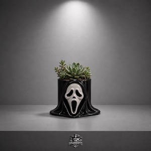 May include: A black ceramic planter featuring the iconic Scream mask design. The planter is filled with green succulents. The mask has a white face with a gaping mouth and black hood. The planter sits on a gray surface.